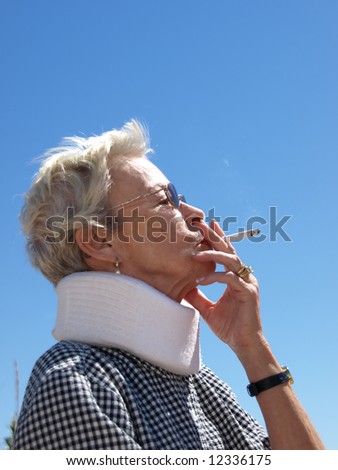 Elderly lady with short-cropped white hair leaning back and taking a drag from a cigarette. She is wearing a neck brace and the photo is set against a clear blue sky