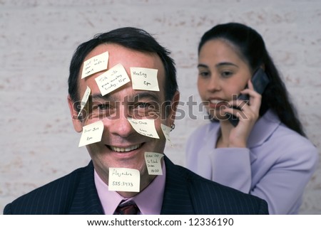 Businessman covered with sticky notes smiling at the camera while a female colleague chats on the phone behind him