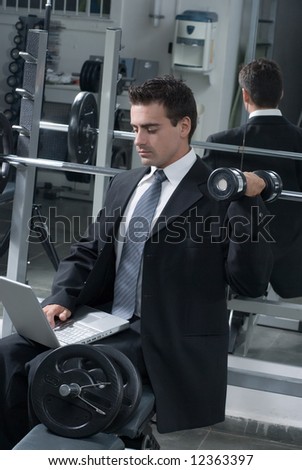 A shot of a businessman, wearing a suit, in gym curling a dumbbell while typing on a laptop.