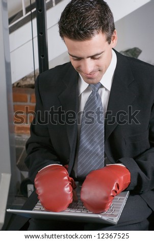 A shot of a businessman, in a suit, typing on laptop while wearing boxing gloves.