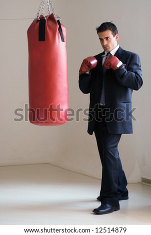 Young attractive businessman in a suit wearing boxing gloves standing ready in front of a heavy punching bag