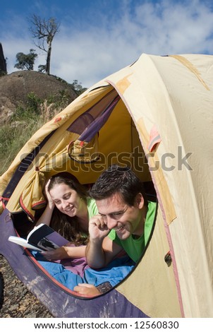 Young smiling couple lying in a tent reading a book and talking on a cellphone.