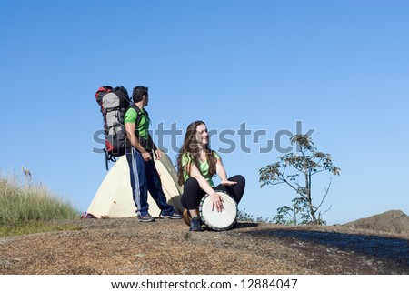 Attractive couple on a camping trip. He has his backpack on and she is playing on a bongo drum. Horizontally framed shot.