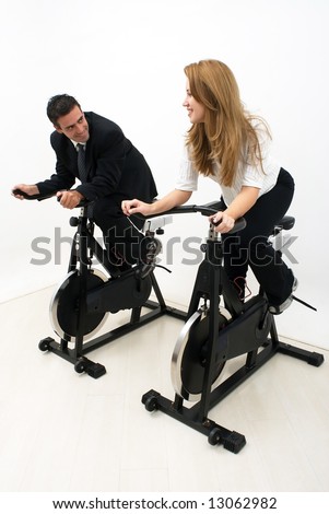 Male and female business colleagues looking at each other and smiling while working out on exercise bikes. Vertical shot isolated against a studio background.