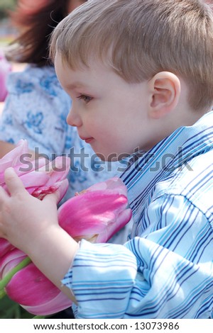 A side shot of a young boy in a blue shirt smelling flowers.