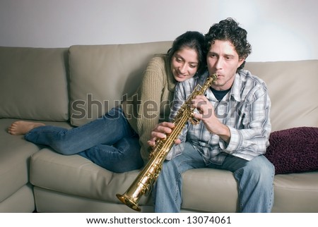 Cute young couple in their living room sharing a tender moment. She is hugging him while he is playing a soprano saxophone