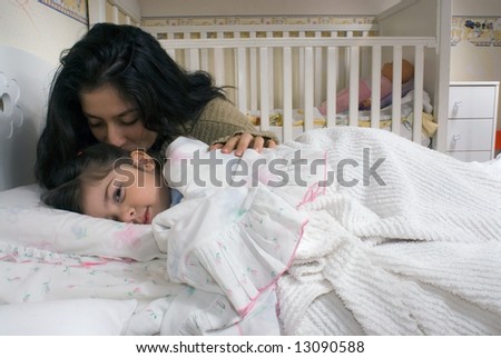 Little girl pretending to be asleep but looking at the camera with a mischievous eye as her mother gives her a goodnight kiss
