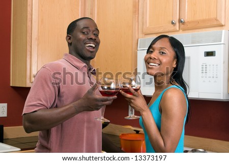 Attractive laughing young African American couple standing in a kitchen clinking wine glasses, in a toast.  Horizontally framed, close cropped, shot with the man and woman looking at the camera.