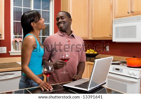 Attractive African American couple, laughing at each other, standing in a kitchen, while using a laptop.  Horizontally framed shot with the man and woman looking at each other, laughing.