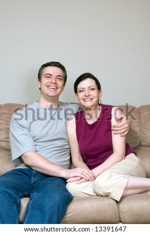 Attractive couple sitting on their living room couch with their arms around each other. Both are smiling at the camera. Vertically framed shot.