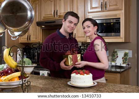 Attractive couple exchanging a gift and sharing a cake in their kitchen. Both are looking at the camera with a neutral expression. Horizontally framed shot.