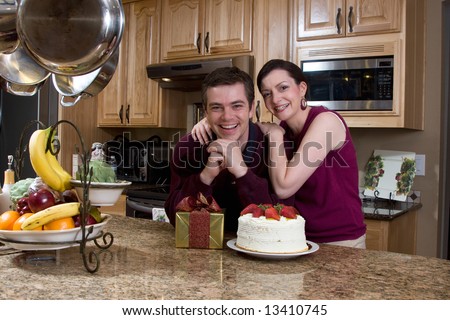 Attractive couple leaning on their kitchen counter which is covered with a cake and a present. They have a huge smile on their faces and are looking at the camera. Horizontally framed shot.