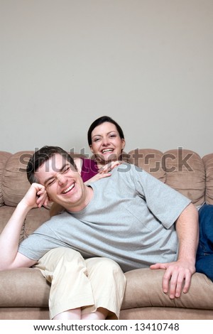 Couple lounging together on their living room couch. He is lying in her lap. Both are laughing at the camera. Vertically framed shot.