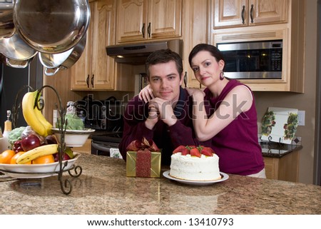 Attractive couple leaning on their kitchen counter which is covered with a cake and a present. They have a neutral expression on their faces and are looking at the camera. Horizontally framed shot.