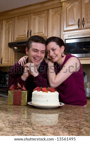 Attractive couple leaning on their kitchen counter which is covered with a cake and a present. They are laughing heartily and looking directly at each other. Vertically framed shot.