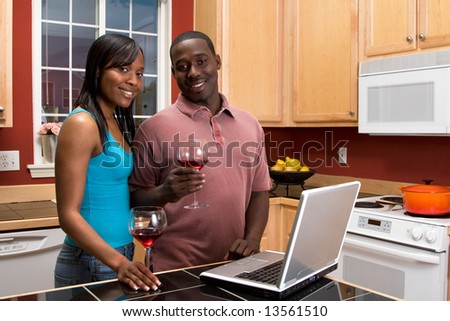 Attractive smiling African American couple standing in a kitchen, holding wine glasses, while using a laptop. Horizontally framed shot with the man and woman looking at the camera.