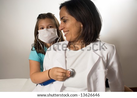A young girl wearing a doctor's mask listens to the doctor's heartbeat with a stethoscope while the doctor sits on an examining table. Horizontally framed photograph