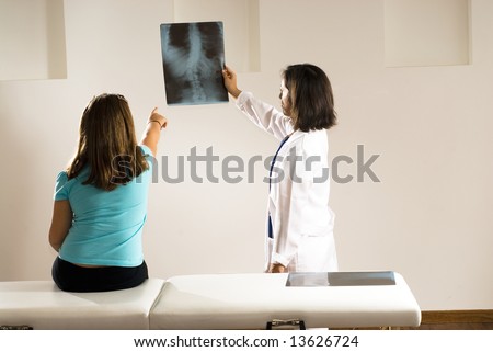 Female doctor holds up an x-ray while a young girl sitting on an examining table points to it. Horizontally framed photograph