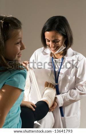 Female Doctor bandages a young girl\'s foot. The doctor\'s mask is partially on her face and you can see she is smiling while she works. The girl is also smiling. Vertically framed photograph