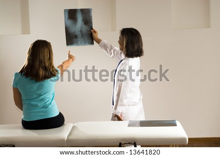 Girl pointing to an x-ray of her back while the smiling doctor holds it up to the wall for her to see. Horizontally framed photograph