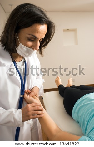 A female doctor wearing a mask checks a patient\'s blood pressure. The patient is lying down on an examining table barefoot. Vertically framed photograph