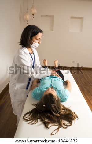 Female doctor wearing a mask checks the abdomen of a barefoot young girl lying down on an examining table. Vertically framed photograph