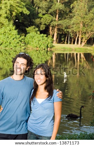 Happy, smiling couple embrace as they stand in front of a wooded pond with swans in it. Vertically framed photograph.