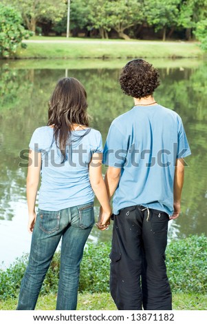 Couple holding hands facing away from the camera looking out at a pond. Vertically framed photograph.