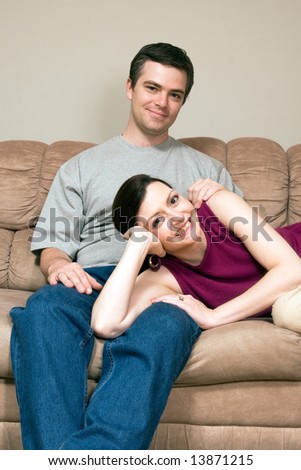 Smiling, happy couple sitting on a couch. They are smiling and he has his arm around her as she lays across his lap. Vertically framed photograph