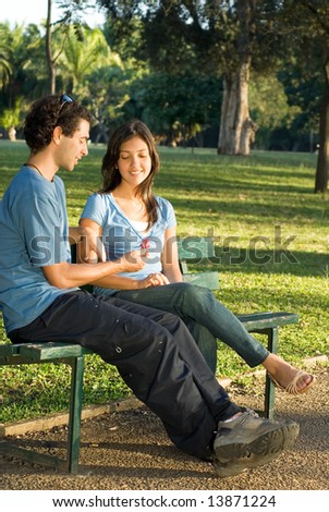Young couple sitting on a park bench. They are smiling at each other as he shows her a red flower. Vertically framed photograph