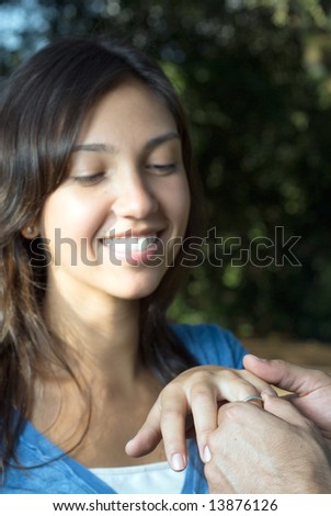 A woman smiles as someone slips an engagement ring upon her finger. Vertically framed photograph.