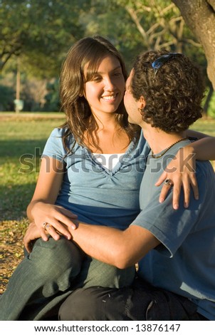 Happy, couple sit in the woods. She sits on his lap a they look at each other and smile. Vertically framed photograph