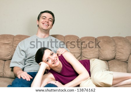 Smiling, happy couple sitting on a couch. They are smiling and he has his arm around her as she lays across his lap. Horizontally framed photograph