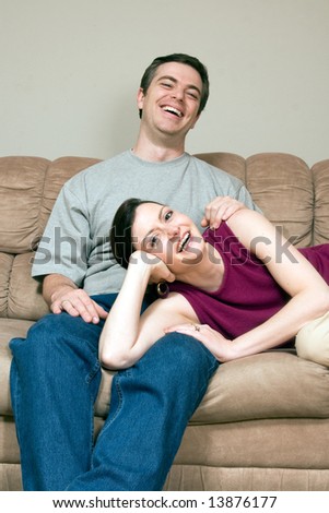 Smiling, happy couple sitting on a couch. They are laughing and he has his arm around her as she lays across his lap. Vertically framed photograph