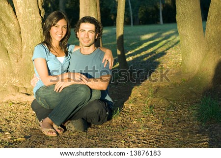 Happy, attractive couple sit in the woods. She sits on his lap a they both smile. Horizontally framed photograph
