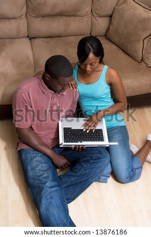 Couple sitting on the floor in front of a couch. They are looking at a laptop computer together. She has her arm on his shoulder. Vertically framed photograph
