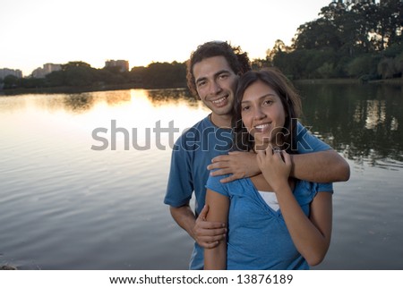 A happy couple embraces as they stand in front of a pond at sunset. Horizontally framed photograph.