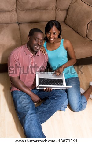 Happy, attractive, couple sitting on the floor in front of a couch. They are holding  a laptop computer together and both are smiling at the camera. Vertically framed photograph
