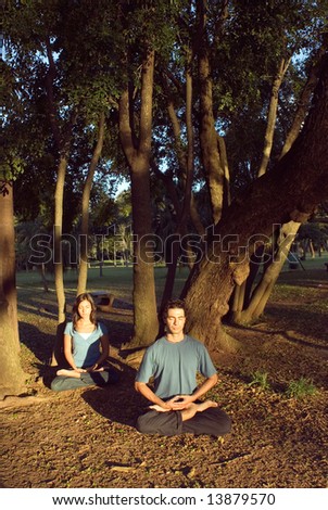 A man and woman practice yoga in a park. They sit cross legged with their eyes closed and are surrounded by a peaceful setting of beautiful trees. Vertically framed photograph