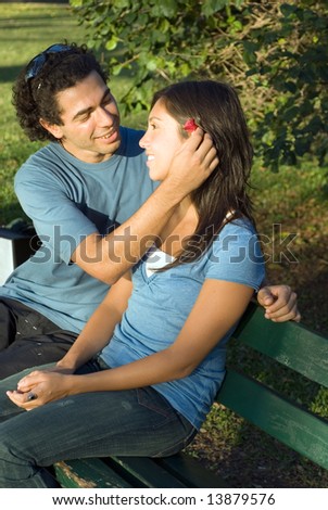 Happy couple on a park bench look at each other as he places a red flower behind her ear. Vertically framed photograph.