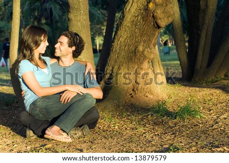 Happy, couple sit in the woods. She sits on his lap a they look at each other and smile. Horizontally framed photograph