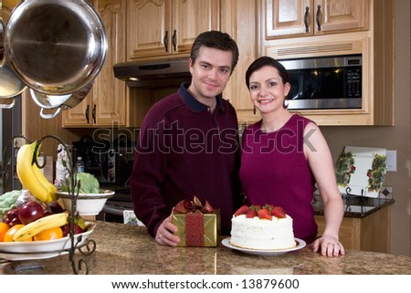 Happy couple in a kitchen with a neutral expression on their faces. He is holding a present. Horizontally framed photograph.