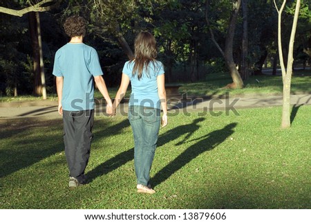 Young couple walking through a park holding hands on a sunny day. They are facing away and you can see their shadows. Horizontally framed photograph