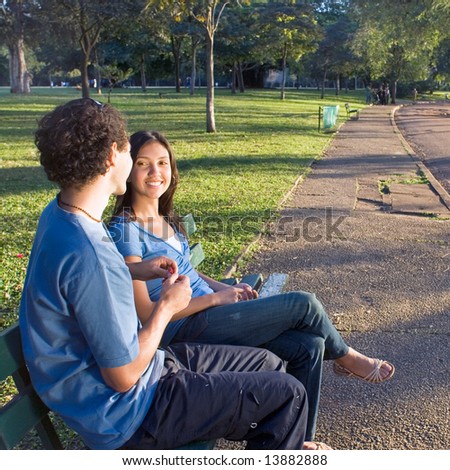 Young couple sitting on a park bench. They are smiling at each other as he shows her a red flower. Horizontally framed photograph