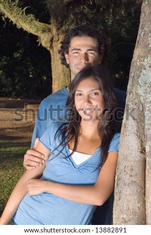 Portrait of a happy couple leaning against a tree in a park. They are smiling as he holds her arm. Vertically framed photograph.