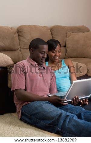 Happy, attractive, couple sitting on the floor in front of a couch. They are looking at  a laptop computer together. She has her arm on his shoulder. Vertically framed photograph
