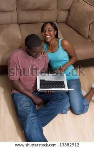 Couple sitting on the floor in front of a couch. They are looking at a laptop computer together. She has her arm on his shoulder and is laughing. Vertically framed photograph