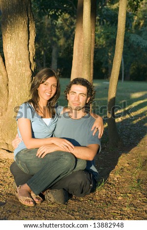Happy couple sitting on the ground under the trees. She is sitting on his lap and they are smiling. Vertically framed photograph.