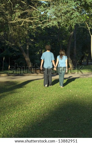 Young couple walking through a park holding hands on a sunny day. They are facing away and you can see their shadows. Vertically framed photograph