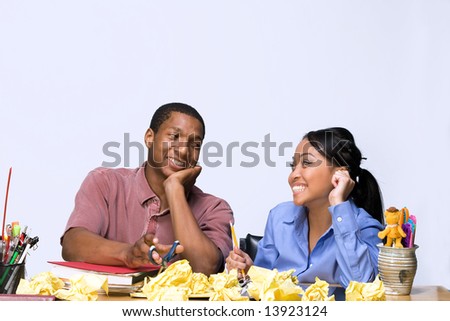 Two Teens are are seated at a desk looking at each other. There are  folders, pens, pencils, and crumpled paper on the desk. Horizontally framed photograph